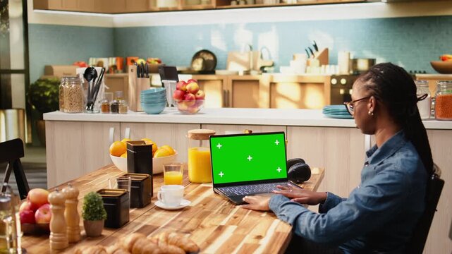 Isolated green screen laptop mockup with woman at kitchen table, using chroma key screen replacement on computer while teleworking stress-free indoors at home with wireless digital communication.