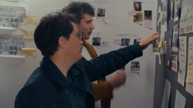 Two male filmmakers examining visual references moodboard on wall and discussing script details during preproduction planning session in creative studio
