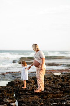 Expecting mother and daughter exploring rocky shore during sunset at the beach