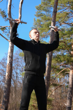 Young adult man performs strength training with adjustable metal dumbbells resting on shoulders during outdoor workout in pine forest. Black sportswear, standing pose, healthy active lifestyle fitness