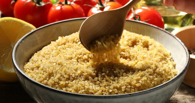 Woman with raw bulgur in bowl and spoon at wooden table, closeup