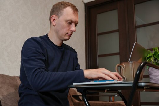 Adult man writes in notebook while looking at laptop in home workspace. Calm concentrated expression accompanies organized desk with computer pen and coffee mug. Concept of remote work, online