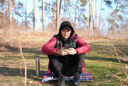 Young adult man sits on picnic blanket in pine forest while using smartphone. Casual outdoor clothing, relaxed posture mobile communication, digital lifestyle travel break, remote connection copy