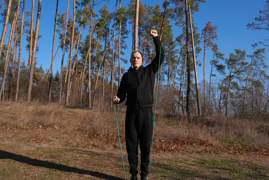Young adult man performs resistance band exercise during outdoor fitness session in pine forest. Elastic training band black athletic clothing, healthy lifestyle, functional strength endurance