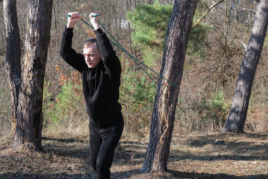 Young adult man performs resistance band exercise during outdoor fitness session in pine forest. Elastic training band black athletic clothing, healthy lifestyle, functional strength endurance