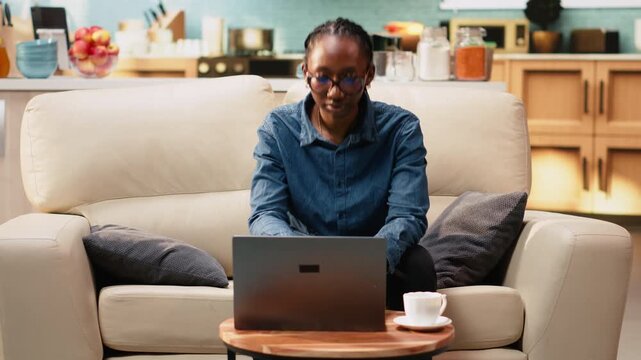 Black woman working remotely from home setup and drinking coffee, focused on solving daily administrative tasks using laptop and internet. Digital nomad lifestyle, flexible work routine.