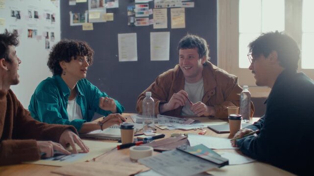 Creative team of four cheerful film crew members smiling and sharing ideas during preproduction meeting around office table with clapperboard and papers on it
