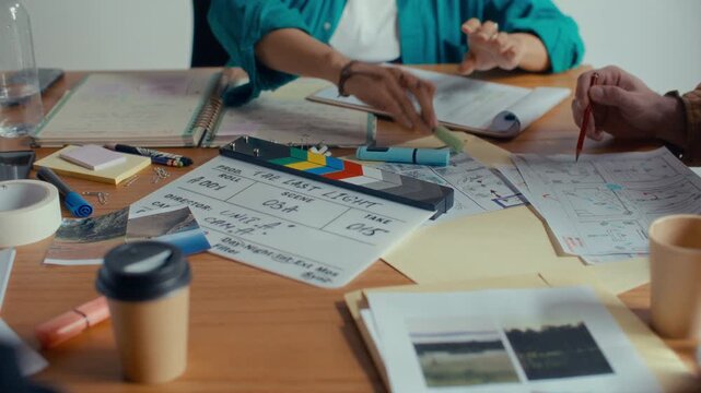 Creative film crew reviewing location reference photos, storyboard and script during discussion at meeting table with clapperboard and printed materials on it. Close up view