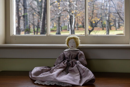 A vintage cloth doll with a painted face, called a rag or cloth folk doll, in a pink dress with puffy sleeves, a lace collar, and a petticoat. The doll's hair is blonde. The toy sits on a wooden floor