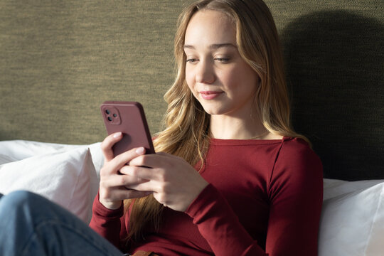 Adolescent female on bed leaning against olive headboard in maroon top and jeans holding rose phone
