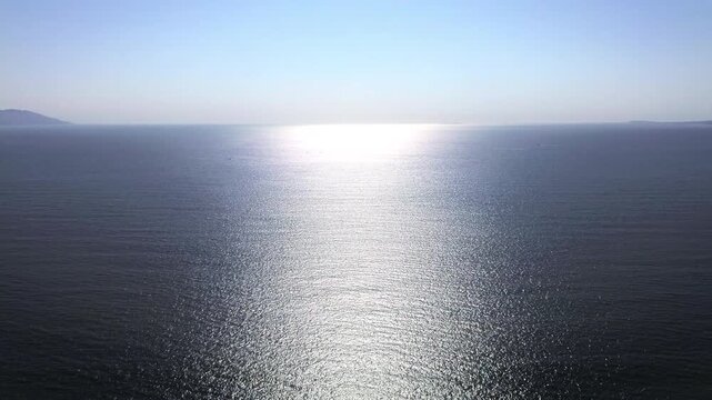 Magnificent sea horizon view during sunrise with bright sun glare reflecting on calm blue water surface as drone moves forward in peaceful clear sky environment