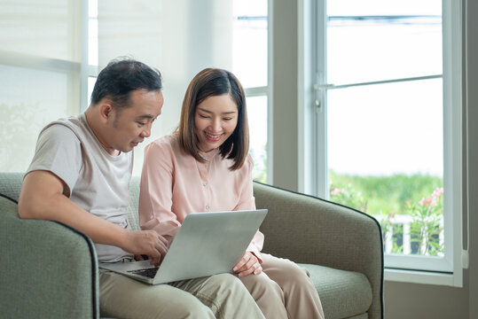 Smiling Asian husband and wife using laptop computer together on sofa near window at home. Online shopping, financial planning, and lifestyle concept. Human-created, not AI.