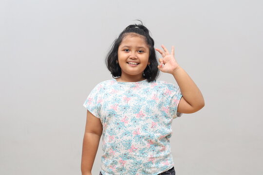 Smiling Asian schoolgirl showing an OK hand gesture isolated on gray background.