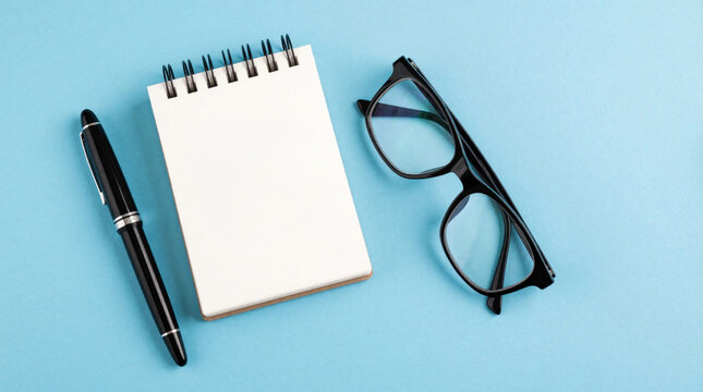 Blank spiral notepad with black pen, eyeglasses on blue background. Minimalist workspace supplies for business planning note taking. Top view of stationery set for office work, study, creative idea