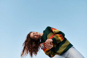 Plakat Young woman in a colorful striped sweater smiles while taking a selfie with her phone against a blue sky. Wind blows her long hair. Concept of joy, communication, and mobile lifestyle.