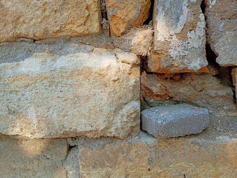 A detailed macro texture of a damaged ancient stone wall made of large rough limestone coquina blocks with a loose grey concrete brick fragment inside a gap, 