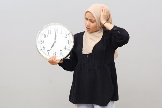 Stressed Asian Muslim woman in hijab holding a wall clock and touching her head, worried about a deadline, isolated on gray background.
