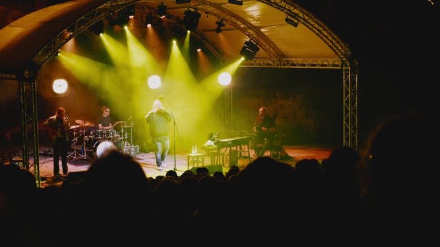 Wide view, live band performing on stage under dramatic yellow concert lighting, with musicians playing before a silhouetted audience at an evening show