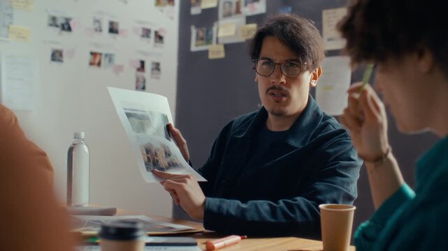 Focused film director pointing to printed location scouting reference images, explaining scene environments to his creative crew during preproduction meeting
