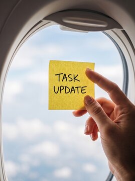 Hand holding yellow sticky note with task update text against airplane window, business travel and remote work concept.