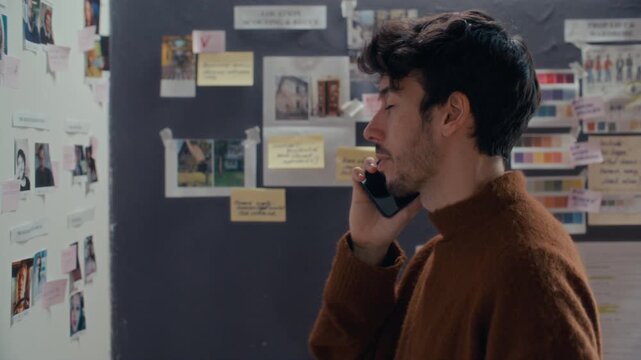 Young filmmaker talking on mobile phone, discussing casting photos and location scouting images pinned to moodboard wall in creative studio