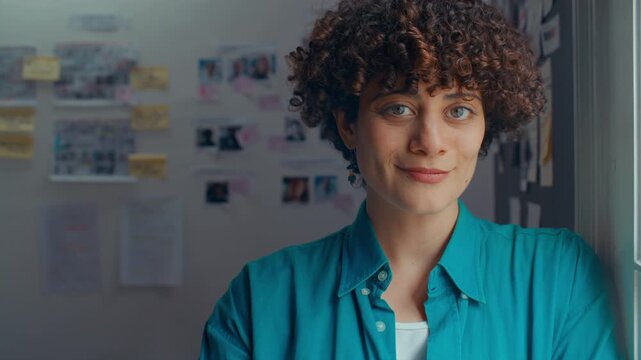 Young beautiful female film producer with curly hair posing confidently for camera beside window in creative studio, with casting boards and storyboards in background. Video portrait