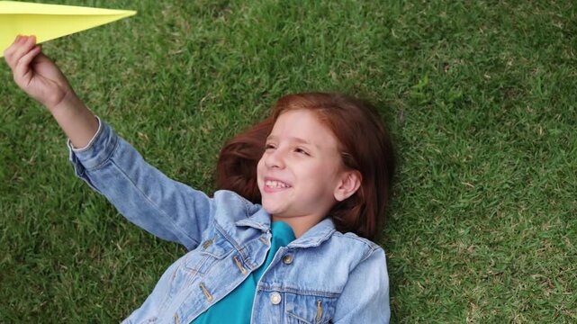 Happy redhead girl playing with paper airplane toy on grass outdoor - Childhood imagination and creativity concept - Top view