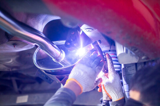 Selective focus to hands of mechanic Argon TIG Welding Exhaust Pipe Underneath Car in Auto Workshop. Welding stainless steel exhaust pipe system under a vehicle with bright sparks in service garage.