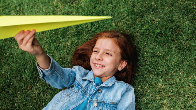 Happy redhead girl playing with paper airplane toy on grass outdoor - Childhood imagination and creativity concept - Top view