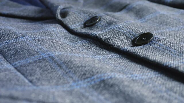A close-up of a grey wool tweed blazer featuring a blue plaid pattern and dark buttons, highlighting the refined fabric texture and classic masculine tailoring details.