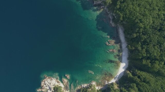 Clear emerald shallows meet a curved pebble beach beside dense coastal forest with submerged rocks and a bright stone outcrop visible along the shore during a vertical aerial glide above the cove