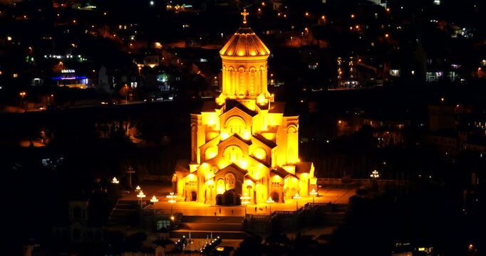 Holy Trinity Cathedral rises brightly illuminated above dark Tbilisi neighborhoods at night with its tall central dome and cross standing against smaller urban lights