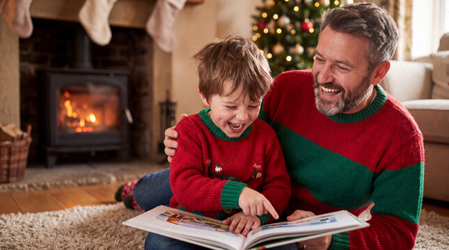 Cozy Caucasian father and son in matching sweaters laughing while reading a Christmas book by the fireplace, perfect for holiday evening visuals.