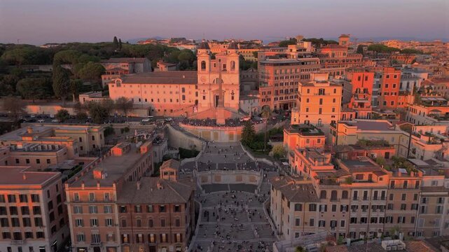 Aerial drone shot in Rome Italy captures iconic Spanish Steps and Trinita dei Monti church bathed in warm golden hour light with tourists gathering on famous stairway surrounded by historic buildings.