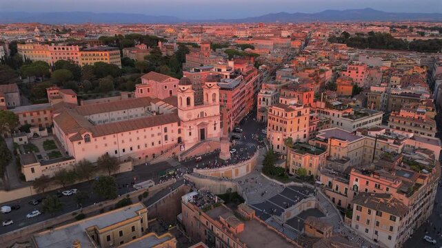 Aerial drone shot in Rome Italy capturing the iconic Spanish Steps and Trinita dei Monti church bathed in golden sunset light with historic terracotta rooftops extending across the cityscape.