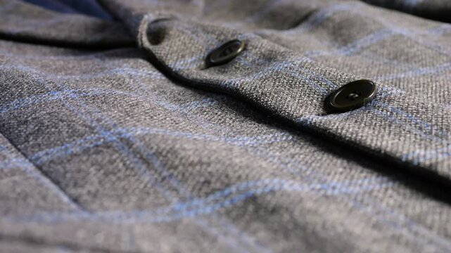 A close-up of a grey wool tweed blazer featuring a blue plaid pattern and dark buttons, highlighting the refined fabric texture and classic masculine tailoring details.