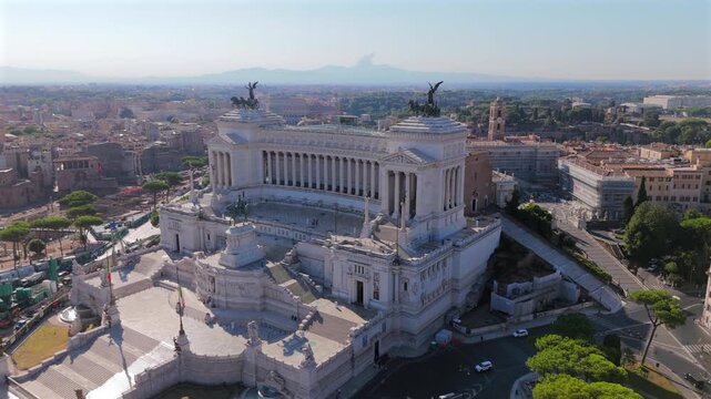 Aerial drone shot in Rome capturing Piazza Venezia and magnificent Altare della Patria monument. White marble columns and bronze quadriga statues crown the neoclassical structure against city skyline.