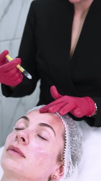Vertical video. Beauty therapy. Facial treatment. Professional specialist in gloves applying product using applicator massaging female forehead for rejuvenation procedure.