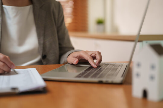 Businesswoman reviewing property loan documents on a laptop beside a house model, representing mortgage financing, banking, real estate investment, lending and home ownership planning.