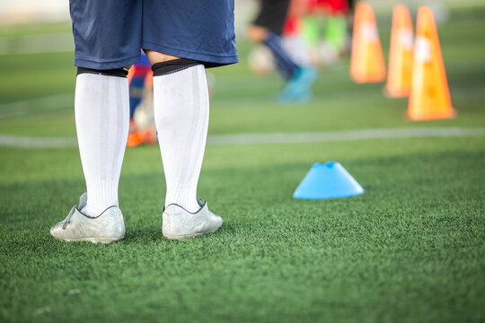 Rear view of a football player wearing white socks and cleats standing on artificial grass pitch during practice with training cones blurred in background.