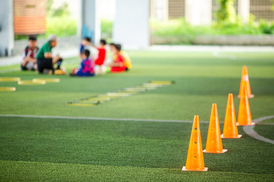 Selective focus to row of bright orange soccer training cones on artificial grass pitch with a blurred background of a coach talking to youth football players during practice.