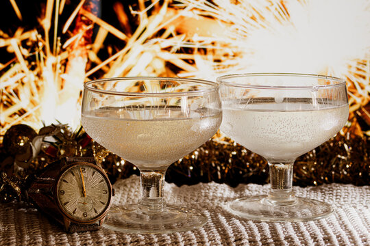 Two coupe glasses of sparkling champagne on a white knitted tablecloth beside a vintage watch at midnight, golden sparklers igniting a cozy, celebratory bokeh filled scene