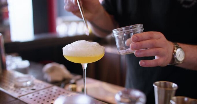 Serving cocktail. Mixologist work. Barkeeper hand tweezers adding dried flower petal to frothy alcoholic yellow liquid on pub counter.