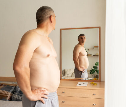 An overweight middle-aged man stands shirtless with his back to camera, looking at his reflection in mirror and assessing changes in his physique. Real person, weight and health monitoring