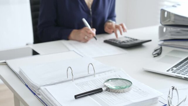 Professional analyzing financial data, writing notes near laptop, magnifying glass, documents, binders on workplace desk. Audit and taxes concept