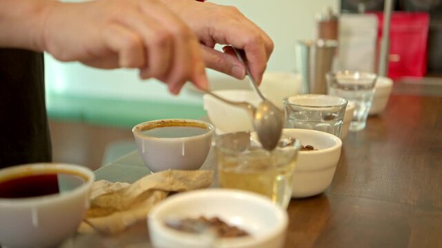 A barista is using a spoon to skim the coffee grounds floating on the surface of the brewed coffee, preparing it for a Q-Grader to taste