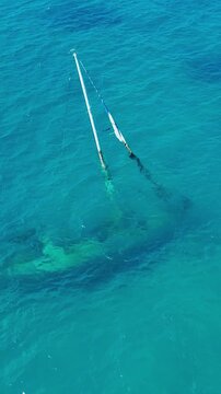 Aerial view sunken yacht in turquoise water, clear visibility revealing leaning mast and halfburied hull, coral patches on seabed, slow current and cinematic drone sweep, survey, vertical video