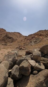 Panning shot from large boulders to a dry, rocky desert mountain under a bright sun.