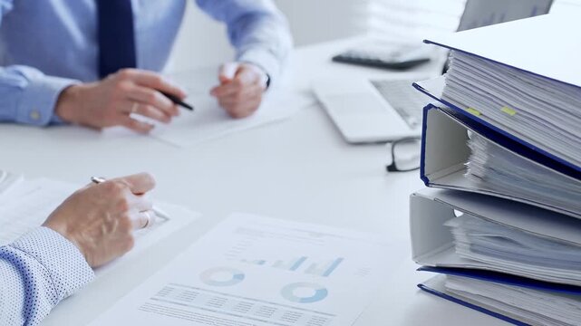 Office workers or auditors are analyzing financial reports and checking documents while sitting at white table with a stack of blue folders in the foreground. Audit and taxes theme in business