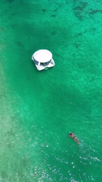 Aerial turquoise sea with lone swimmer, drone captures woman swimming near white floating gazebo over shallow coral, sunlit ripples, tranquil resort vibe, summer travel and leisure,Maldives